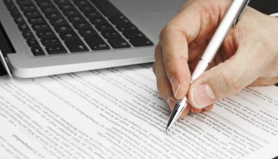 A hand holding a pen next to a laptop and a piece of paper