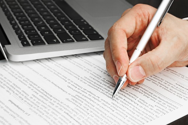 A hand holding a pen next to a laptop and a piece of paper